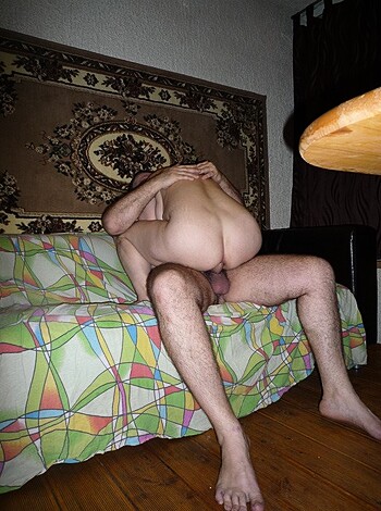 A hairy dude is giving head to some other guy sitting on a couch while their dicks are out.