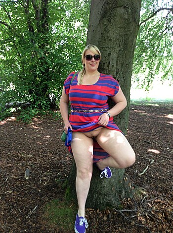 Image of a slim woman with long hair dressed in a red and blue checkered dress sitting on a tree branch while exposing her naked body.