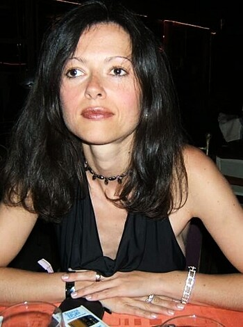 Here's a factual description of the image: A busty brunette bitch is seated at a fuckin' table wearing a tight-assed black dress, smiling seductively while gazing towards her handsome ass suitor.