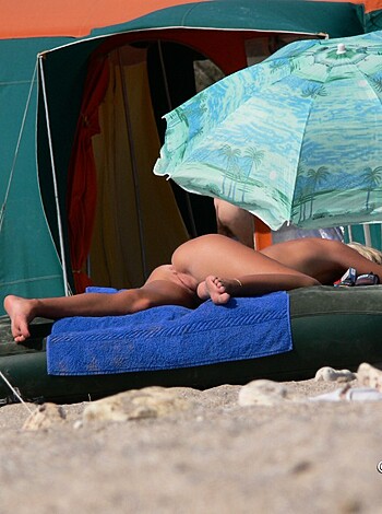 """
Dude on the sandy shore with no shirt on, chilling under a sunshade while some fully-clothed folks lounge around him.
""