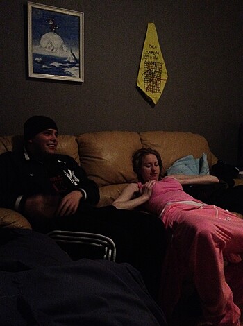 A couple is seen relaxing on a couch, with the guy sporting a stupid grin while the lady's dressed in a pink pair of flannel pajamas.