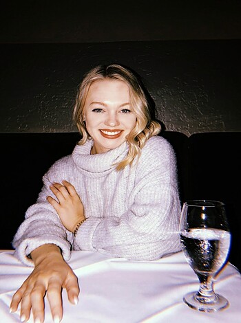 Image depicts busty brunette chick with perfect tits, wearing cozy gray sweater, sitting at table and grinning like a slutty whore, showing off her pearly whites.