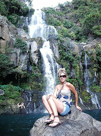 A busty blonde broad is basking in nature, with her wet tits exposed and her ass hanging out as she sits on some rocks near a cascading waterfall.