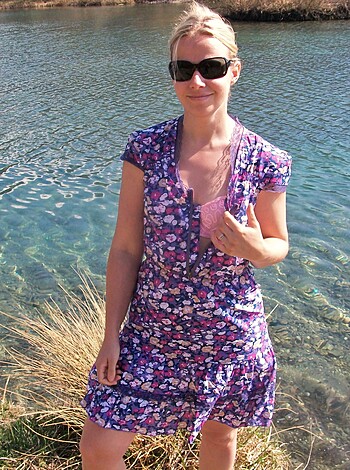A chick with her arm extended, showing off her ring finger while standing next to some water.