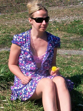 Image of a busty blonde chick in tight jeans and a cropped top, smiling with her tongue out while gripping her pussy while perched on some lawn like a fucking dumbass with a daisy in her hand.