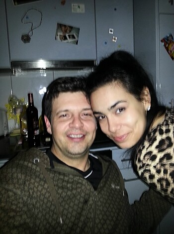 The guy is sitting on a stool and his girlfriend is kneeling on the ground, leaning over him, as they take a sexy photo in their kitchen.