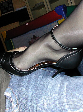 The image depicts a footwear item, belonging to a woman, resting upon the crotch region of a seated male figure.